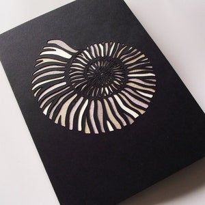 May include: A black card with a white cutout of an ammonite fossil. The ammonite is a spiral shell with intricate details.