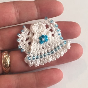 May include: A small crocheted angel ornament with a white crocheted dress and blue beads. The angel has a blue flower on its chest.