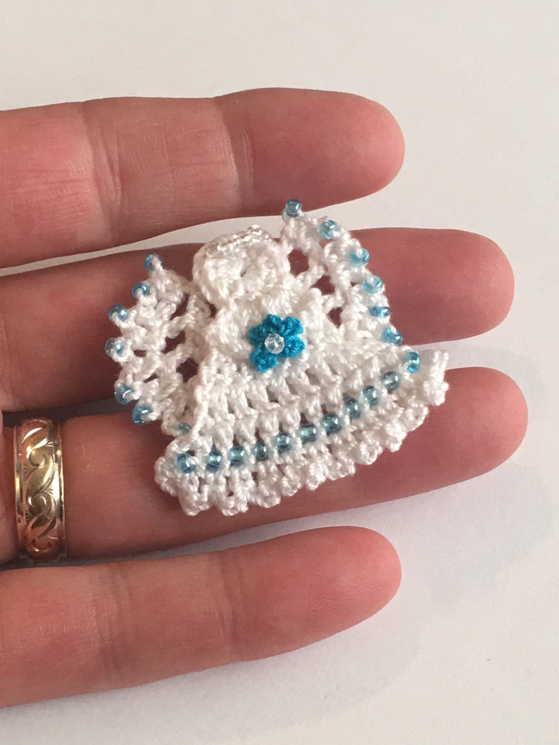 May include: A small crocheted angel ornament with a white crocheted dress and blue beads. The angel has a blue flower on its chest.