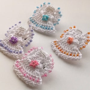 Puede incluir: Cuatro adornos de ángel de ganchillo con ribete de cuentas. Cada ángel es blanco con una flor de color diferente en el centro. Las flores son moradas, azules, naranjas y rosas.