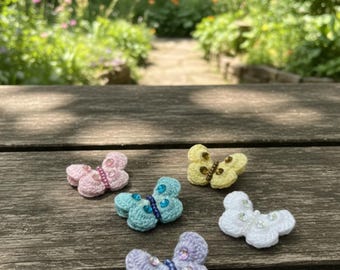 May include: A collection of five crocheted butterfly ornaments in various pastel colors, including pink, blue, yellow, and white. Each butterfly features beaded accents and is displayed on a weathered wooden surface, with a blurred garden path in the background.