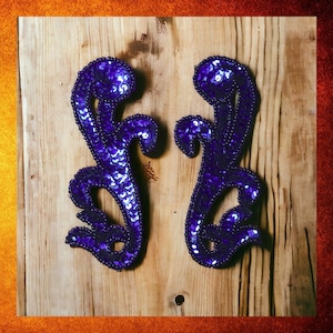 May include: Two purple sequined fabric appliques in the shape of stylized floral designs. The appliques are on a wooden surface.