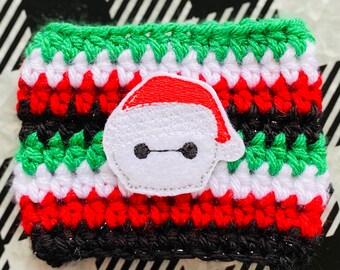 May include: A handmade crochet coffee cup cozy in Christmas colors: green, white, red, and black. The cozy features an embroidered Baymax character wearing a Santa hat. The cozy is designed to fit a standard coffee cup.