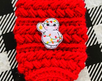 May include: A red crocheted coffee cup cozy with a white embroidered patch of a cartoon character decorated with Christmas lights. The cozy is made of thick, textured yarn.