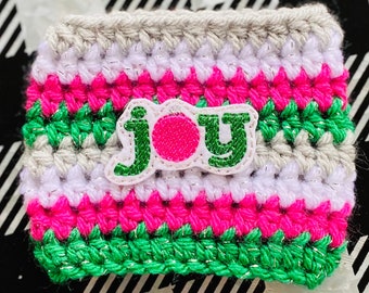 May include: Crocheted coffee cup cozy with a striped pattern in pink, green, white, and gray. The word "JOY" is appliqued on the front in green and pink, with a small ornament detail above the "O".