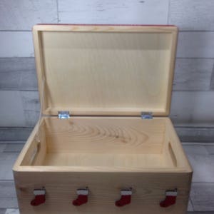 May include: A wooden storage box with a hinged lid, open to reveal the interior. The box is decorated with four red Christmas stocking ornaments on the front. The box is made of light-colored wood.