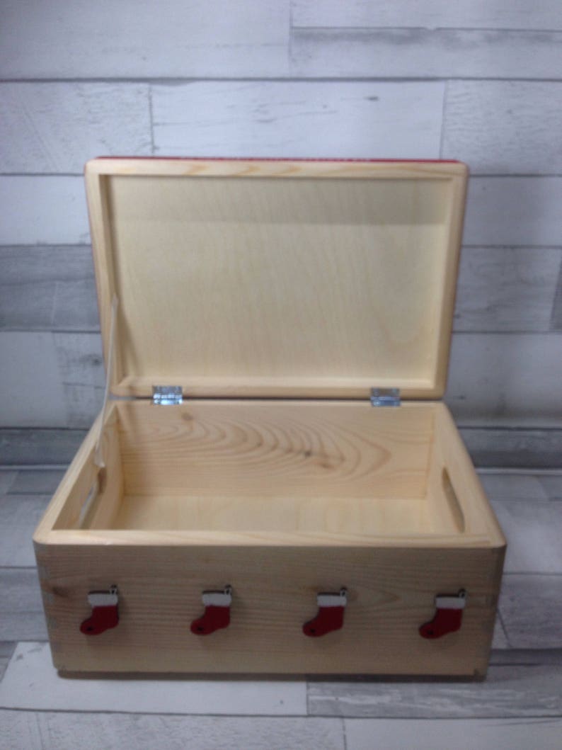 May include: A wooden storage box with a hinged lid, open to reveal the interior. The box is decorated with four red Christmas stocking ornaments on the front. The box is made of light-colored wood.