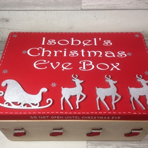 May include: A red Christmas Eve box with white text that reads "Isobel's Christmas Eve Box". The box features a silver sleigh and reindeer design. The box is made of wood and has red stocking-shaped handles.
