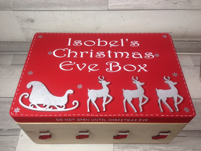 May include: A red Christmas Eve box with white text that reads "Isobel's Christmas Eve Box". The box features a silver sleigh and reindeer design. The box is made of wood and has red stocking-shaped handles.