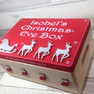 May include: A wooden Christmas Eve box with a red lid featuring white text that reads "Isobel's Christmas Eve Box". The lid has a silver sleigh and reindeer design. The box has red stocking accents.