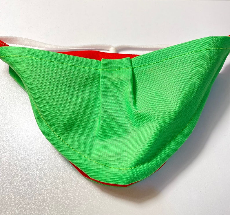 May include: Green and red fabric face mask with a white elastic band. The mask has a pleated design and is sewn with a contrasting thread.