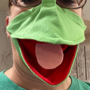 May include: A green felt puppet mask with a red mouth and a pink tongue. The mask is designed to look like a frog.
