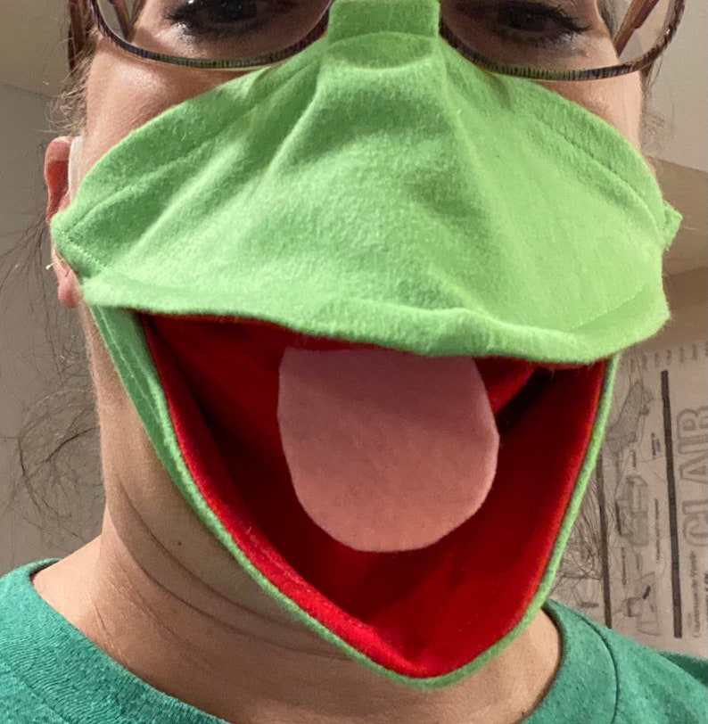May include: A green felt puppet mask with a red mouth and a pink tongue. The mask is designed to look like a frog.