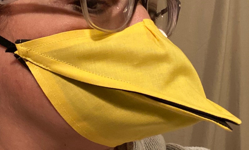 May include: A yellow fabric face mask with a black trim and a beak-like extension. The mask is worn by a person.
