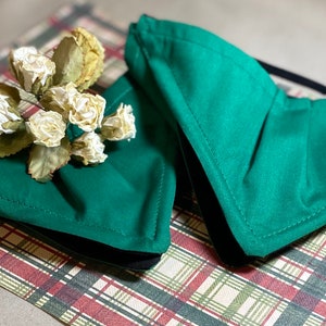 May include: Two green fabric face masks with black elastic ear loops. The masks are folded and lying on a plaid surface.