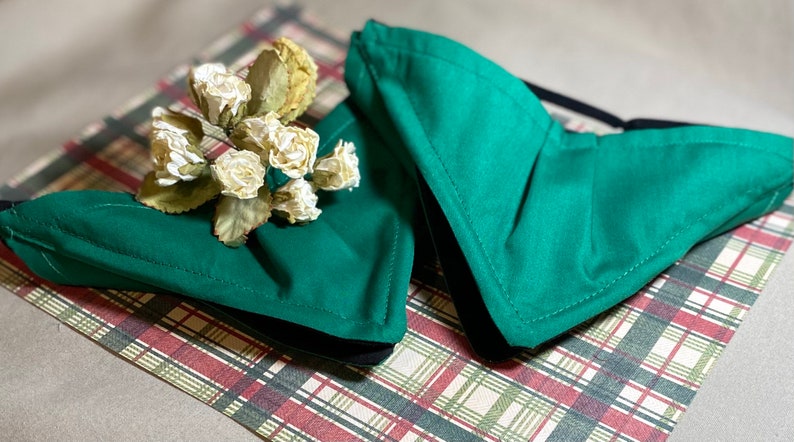May include: Two green fabric face masks with black elastic ear loops. The masks are folded and lying on a plaid surface.