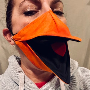 May include: A person wearing a bright orange and black fabric face mask with a large, open beak. The mask is designed to look like a bird's beak.