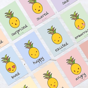 May include: A collection of colorful cards featuring cartoon pineapples expressing different emotions. Each card has a unique pastel background and a word describing the emotion, such as "surprised," "happy," and "angry." The cards are arranged on a light-colored wooden surface.