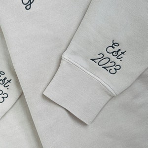 May include: Close-up of a beige sweatshirt sleeve with the embroidered text "Est. 2023" in black script. The cuff is ribbed and the fabric has a soft texture. The sweatshirt appears to be made of a comfortable material.