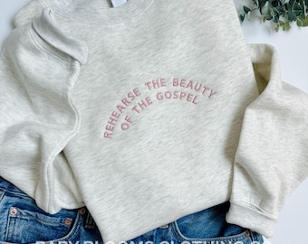 May include: A light gray sweatshirt with the embroidered text "REHEARSE THE BEAUTY OF THE GOSPEL" in pink. The sweatshirt is folded over blue denim jeans. The words "BABY BLOOMS CLOTHING CO." are printed at the bottom of the image.