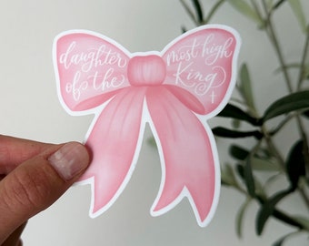 May include: A pink bow-shaped sticker with the words "daughter of the most high King" in white script. The sticker has a white border and is held by a hand.