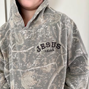 Puede incluir: Una sudadera con capucha con estampado de camuflaje con el texto "JESUS IS KING" en letras negras. La sudadera con capucha presenta una capucha y una paleta de colores neutros de verdes, marrones y grises.