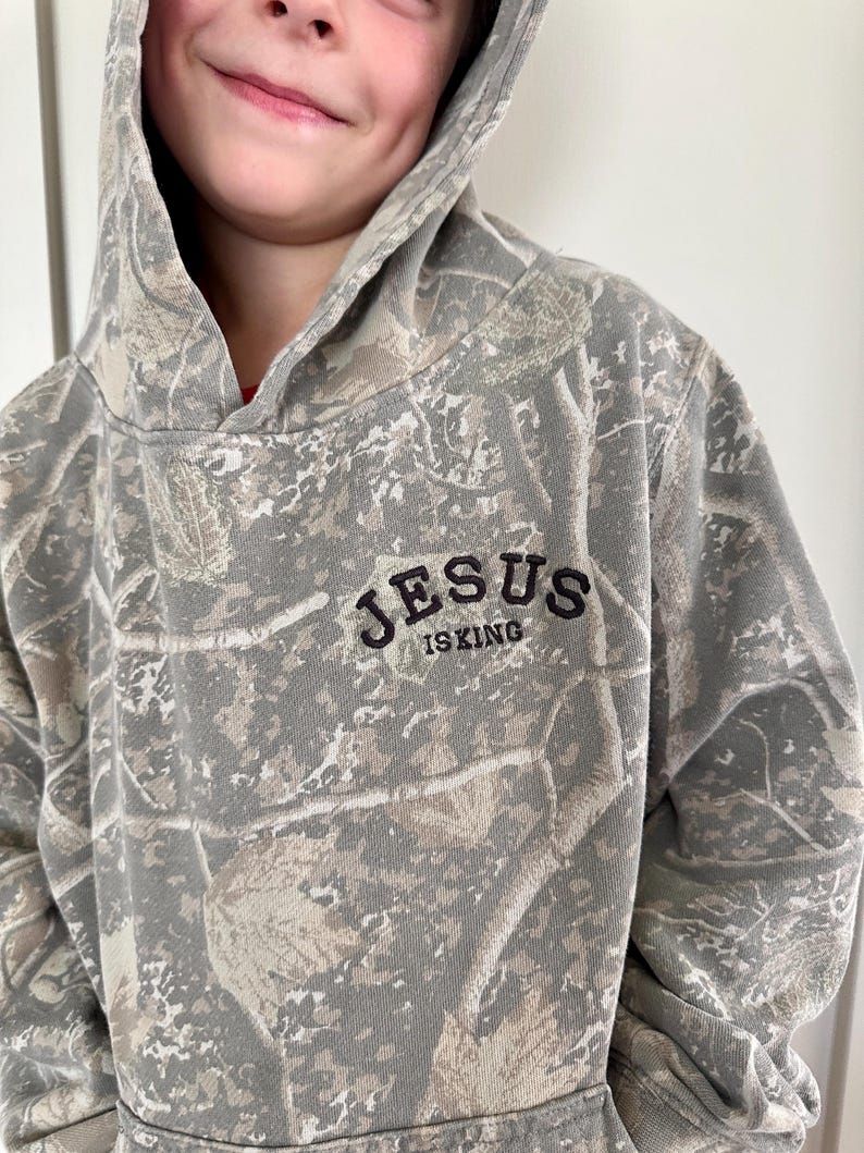 Puede incluir: Una sudadera con capucha con estampado de camuflaje con el texto "JESUS IS KING" en letras negras. La sudadera con capucha presenta una capucha y una paleta de colores neutros de verdes, marrones y grises.