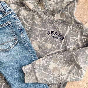 Puede incluir: Una sudadera con capucha con estampado de camuflaje con el texto "JESUS IS KING" bordado en azul marino, combinada con vaqueros de mezclilla azul claro. La sudadera con capucha tiene una capucha con cord&oacute;n y mangas largas. Los vaqueros tienen un dise&ntilde;o cl&aacute;sico de cinco bolsillos.