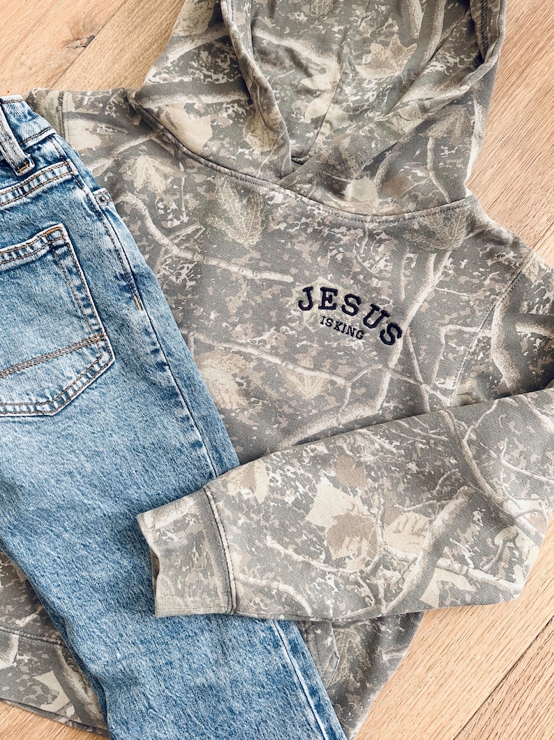 Puede incluir: Una sudadera con capucha con estampado de camuflaje con el texto "JESUS IS KING" bordado en azul marino, combinada con vaqueros de mezclilla azul claro. La sudadera con capucha tiene una capucha con cord&oacute;n y mangas largas. Los vaqueros tienen un dise&ntilde;o cl&aacute;sico de cinco bolsillos.