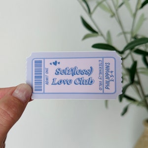 May include: A light blue ticket with the words "Selfless Love Club" in a playful font. The ticket design includes small hearts and the text "Philippians 2:3-4". The ticket is held against a blurred background of greenery.