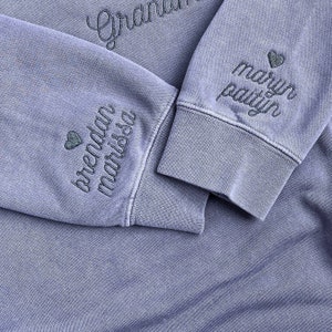 May include: Close-up of a light blue sweatshirt with embroidered text. The word "Grandma" is visible at the top. The sleeves feature names "Brendan Marissa" and "Maryn Paitlyn" with heart accents. The fabric has a faded, vintage look.