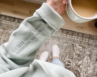 Könnte beinhalten: Hellgrüner Sweatshirt-Ärmel mit dem gestickten Text "BUT TAKE HEART! I HAVE OVERCOME THE WORLD." Eine Person hält eine weiße Tasse mit einem hellbraunen Getränk. Die Person trägt weiß karierte Slip-on-Schuhe.
