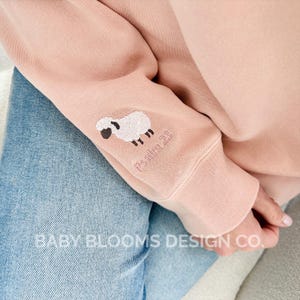 May include: A light pink sweatshirt sleeve with an embroidered sheep and the text "Psalm 23". The sleeve is over a pair of blue jeans. The text "BABY BLOOMS DESIGN CO." is visible at the bottom of the image.