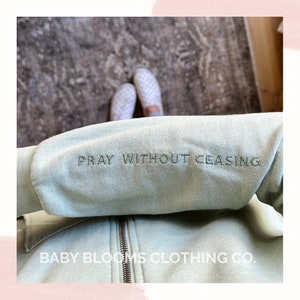 May include: A light green zip-up hoodie with the embroidered text "PRAY WITHOUT CEASING" on the sleeve. The brand name "BABY BLOOMS CLOTHING CO." is printed at the bottom. The hoodie has a silver zipper.