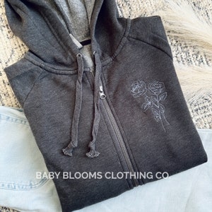 May include: Charcoal gray zip-up hoodie with a drawstring hood and embroidered white rose design. The hoodie features a full zipper closure and the text "BABY BLOOMS CLOTHING CO." at the bottom.