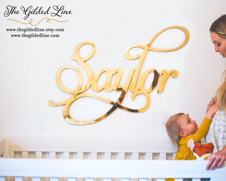 May include: Gold cursive lettering spelling out the name "Sailor" on a white wall. A young child is standing in a crib, reaching up towards the name.