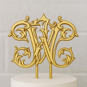 May include: Gold-colored cake topper featuring the intertwined letters "W" and "V" in an ornate, elegant script. The design includes decorative flourishes and is mounted on two slender posts for insertion into a cake. The topper sits atop a white cake.