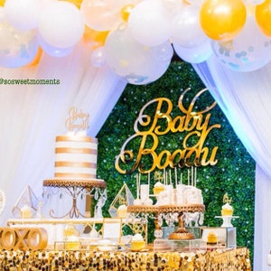 May include: A gold cake with white stripes sits on a cake stand with a gold base and crystal accents. The cake is decorated with a gold topper that says "Baby Bodhi". The cake is surrounded by a gold table cloth and a green backdrop with the words "Baby Bodhi" in gold letters. There are white and gold balloons in the background.