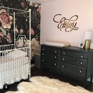 May include: A white crib with a black and white polka dot sheet and a white lace ruffle. The crib is in a nursery with a black and white floral wallpaper, a black dresser with white knobs, and a white rug. The name "Elsie" is written in gold on the wall.