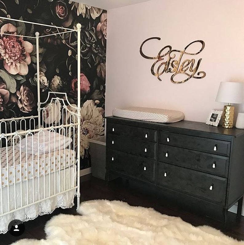 May include: A white crib with a black and white polka dot sheet and a white lace ruffle. The crib is in a nursery with a black and white floral wallpaper, a black dresser with white knobs, and a white rug. The name "Elsie" is written in gold on the wall.