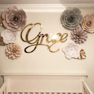 May include: Gold mirror acrylic name sign with the word "Grace" in a cursive font. The sign is surrounded by paper flowers in shades of white, beige, and gray.