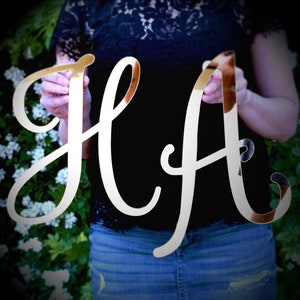 May include: A pair of mirrored gold letters, "HA", are held up against a dark background. The letters are in a cursive font, reflecting the surrounding environment. The letters are likely used for decorative purposes.
