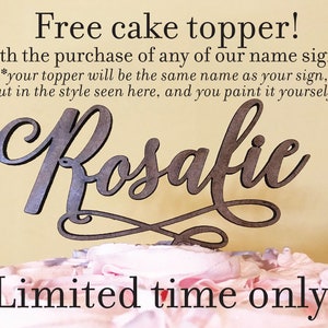 May include: A wooden cake topper with the name "Rosalie" written in a cursive script. The topper is on a pink and white frosted cake. The text "Free cake topper! With the purchase of any of our name signs. *your topper will be the same name as your sign, but in the style seen here, and you paint it yourself! Limited time only!" is written on a yellow background.