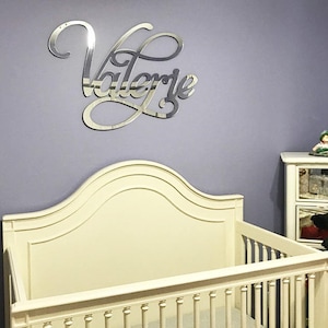 May include: A white crib with a cream-colored headboard and a silver mirror name sign that reads "Valerie" on a light purple wall.