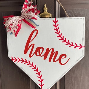 May include: White baseball home plate-shaped sign with red stitching and the word "home" in red script. A decorative bow with baseball-themed ribbon and a jute rope for hanging complete the design.