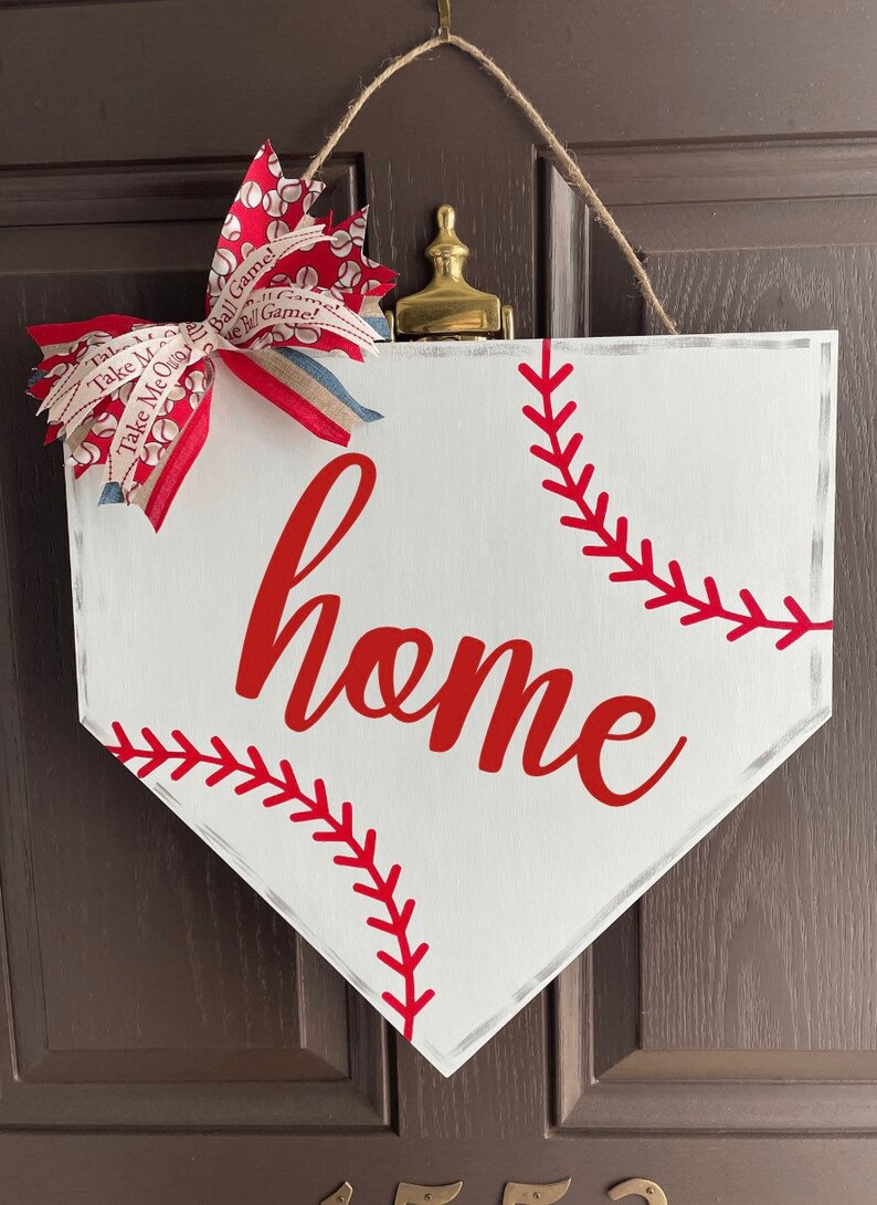 May include: White baseball home plate-shaped sign with red stitching and the word "home" in red script. A decorative bow with baseball-themed ribbon and a jute rope for hanging complete the design.