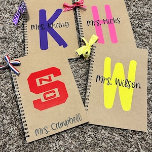 May include: Four brown spiral notebooks with different colored letters and names on the front. The names are "Mrs. Khang", "Mrs. Hicks", "Mrs. Campbell", and "Mrs. Wilson".