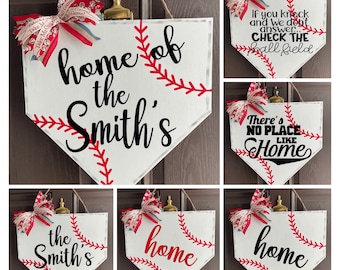 May include: Baseball-themed home decor sign in the shape of a home plate. The white sign features red stitching, a decorative bow, and the text "home of the Smith's". Other signs say "If you knock and we don't answer... CHECK THE ball field" and "There's NO PLACE LIKE Home".