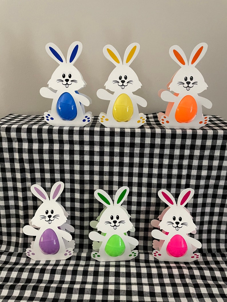 May include: Six white bunny-shaped cardboard holders with colorful plastic eggs inside. The bunnies are standing on a black and white checkered surface.