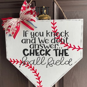 May include: White baseball home plate-shaped sign with red stitching and the text "If you knock and We don't answer... CHECK THE ball field." A red, white, and blue bow is attached to the top. A baseball-themed decorative item.